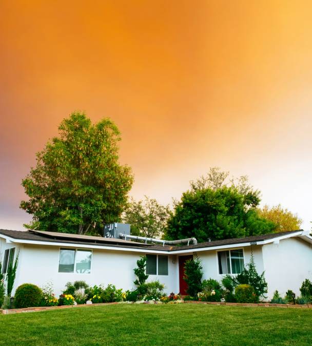 White single-story house with red door surrounded by lush green trees, shrubs, and manicured lawn under an orange gradient sky.