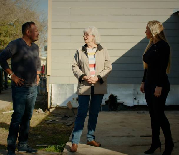 Three people, a man and two women, stand outside a house having a discussion. The man is casually dressed while the women wear jackets.